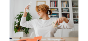 A woman sits at a table, playfully holding up several bills. She appears happy and possibly successful in managing her finances.