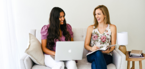 Two women sit on a light gray couch, smiling and working together. One uses a laptop, the other a notebook. A small side table with a lamp and books is beside them.