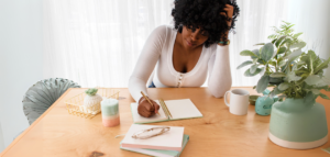 A woman sits at a light wood table, looking stressed as she writes in a notebook. A mug, plants, and other small items are on the table.