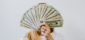 A woman's hand, holds up a fan of US dollar bills, covering her face.