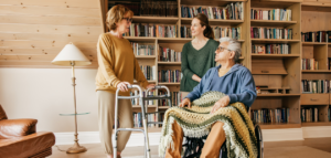 An older man in a wheelchair is surrounded by two women, one standing and using a walker, the other standing beside him. They are in a home library with bookshelves in the background.

