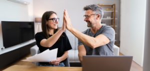 A couple sit at a table, smiling and giving each other a high five. A laptop and papers are on the table.