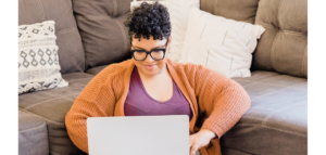 A woman sits on a couch, smiling as she works on a laptop computer.