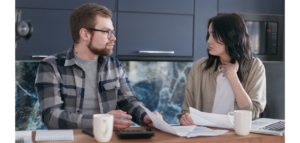 A man and a woman sit at a kitchen table, reviewing finances and looking concerned. A laptop and mugs are also on the table.