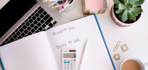 A flat lay image of a workspace showing a laptop, notebook with handwritten personal and business goals, calculator, pencils, sticky notes, paper clips, a succulent plant, and a cup of coffee.