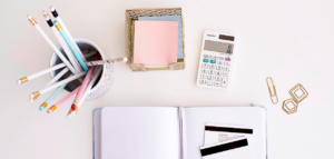 A flat lay photo of a workspace showing a notebook, a calculator, a container of pencils, sticky notes, gold paper clips, and credit cards.
