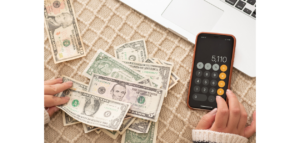 A person's hands sorting through US dollar bills while using a calculator app on their smartphone. 
