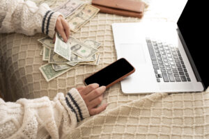 Close-up of a person's hands holding several US dollar bills on a beige blanket, next to a laptop and a smartphone.