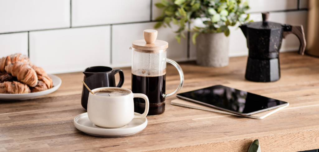 A cup of coffee sits on a wooden counter next to a French press, a small creamer, a plate of croissants, and a tablet.