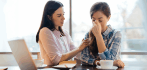 One woman comforting another who appears stressed, with a laptop and coffee cups on the table between them.