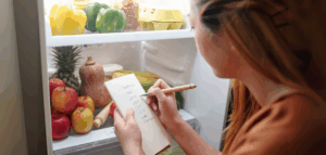 Woman standing in front of an open refrigerator, writing a grocery list on a small notepad with fruits and vegetables visible inside.
