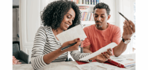 Smiling couple sitting at a kitchen table, reviewing paperwork and notes together, appearing to work on their finances or budget.