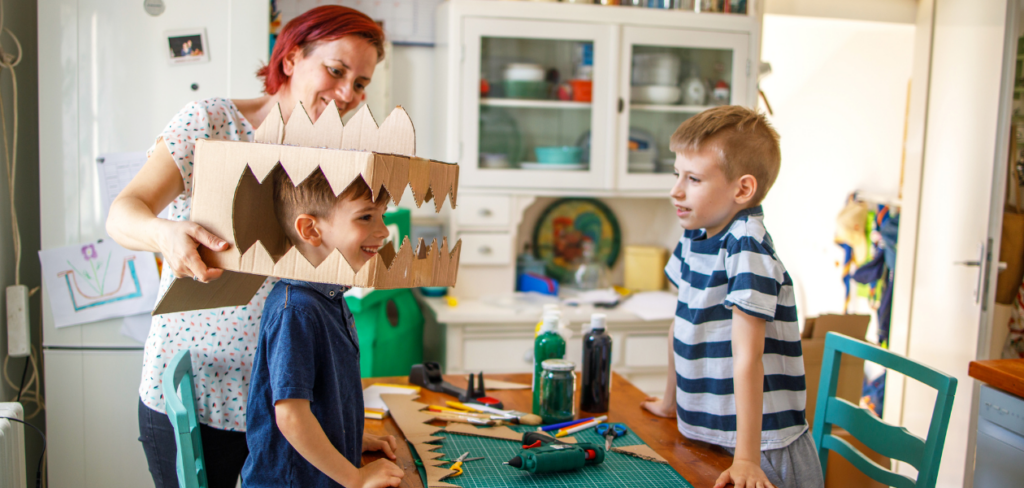 A mother helps her young son put on a cardboard dinosaur mask. His brother watches from the side as they work on a costume project at a kitchen table.