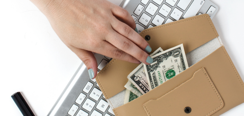 A woman's hand reaches into a wallet next to a computer keyboard, retrieving a few dollar bills.
