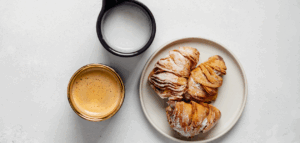 A small plate with two pastries, a cup of espresso, and a small pitcher of milk on a light gray surface.