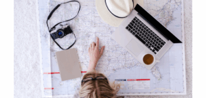 Overhead view of a woman on the floor, planning a trip. A large map of the United States is spread out, and she's pointing to a location. A laptop, camera, notebook, hat, and cup of tea are also arranged on the map.
