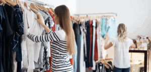 A woman browses clothing racks in a clothing boutique. Another woman is visible in the background, also shopping.