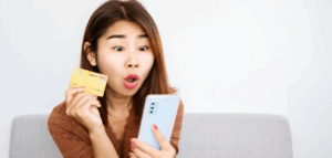 A woman looks surprised as she holds a credit card and a smartphone, seemingly in the middle of an online purchase.