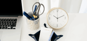 Workspace with a laptop, gold clock, pen holder with pens and scissors, and a small leather notepad holder with a pen.