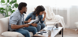 A couple sits on a couch, reviewing bills and looking stressed. A laptop, calculator, and other financial documents are on the coffee table in front of them.