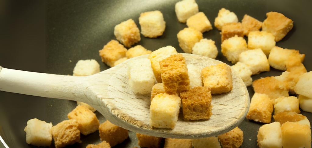 A wooden spoon scoops up  croutons from a dark pan.