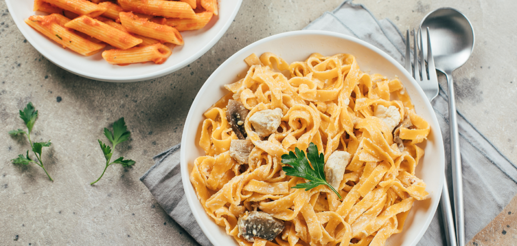 A plate of creamy pasta, alongside a smaller plate of pasta in tomato sauce. A fork and spoon rest on a gray napkin next to the main dish.