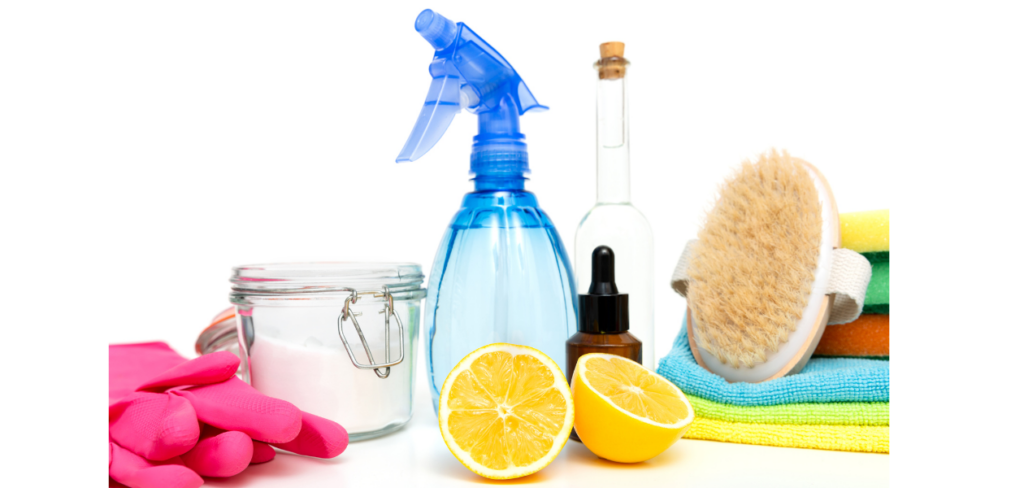 A collection of natural cleaning supplies, a wooden brush, lemon halves, and pink cleaning gloves, all arranged on a white background with colorful cleaning cloths.