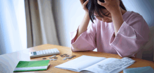 A woman sits at a table, head in hands, looking stressed. A calculator, coins, a notebook, and receipts are on the table in front of her.