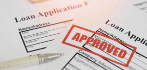 A close-up shot of two loan applications, one of which is stamped "APPROVED" in large red letters. 