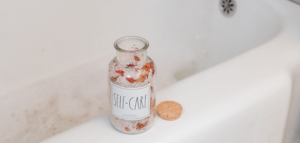A glass jar of bath salts sits on the edge of a bathtub. The jar has a label that reads "SELF-CARE" and the cork lid is next to it.