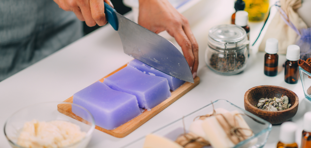 Close-up view of hands using a large knife to cut a block of lavender-colored soap into smaller bars.