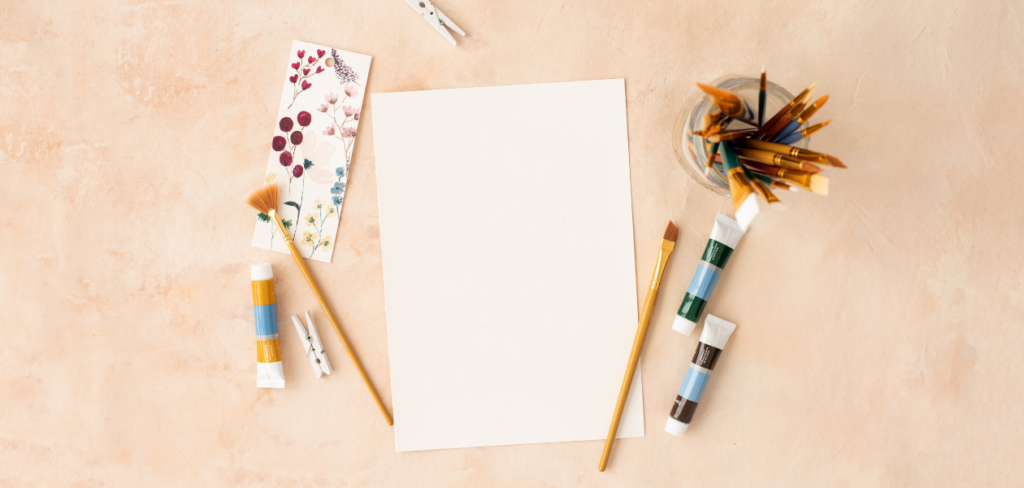 A flat lay image shows a blank sheet of white paper surrounded by art supplies like tubes of paint, paintbrushes, and a floral bookmark.