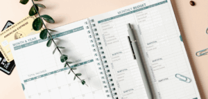 An image of an open budgeting notebook with sections for monthly income, expenses, and savings. Credit cards, a pen, and paper clips are arranged around the notebook.