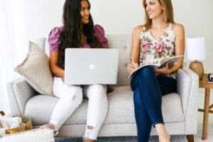 Two women sitting on a couch, one using a laptop and the other writing in a notebook, engaged in conversation.