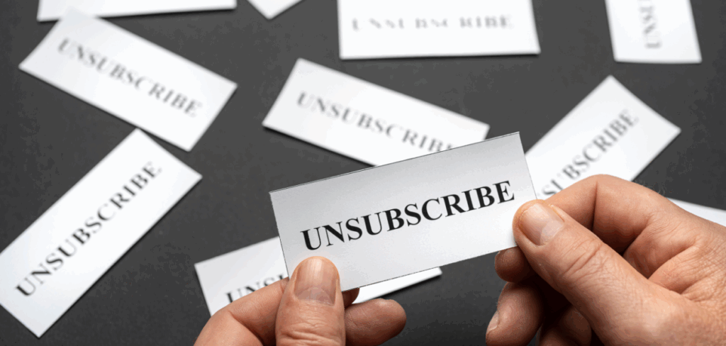 Close-up of hands holding a white card that says “UNSUBSCRIBE,” with more similar cards scattered on a dark background.