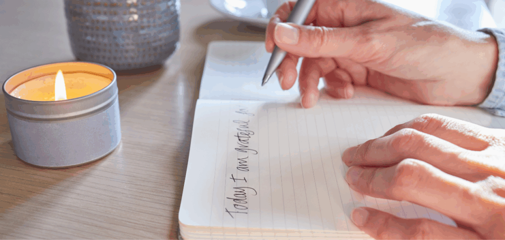 Close-up of hands writing "Today I am grateful for" in a notebook, with a lit candle in the background.